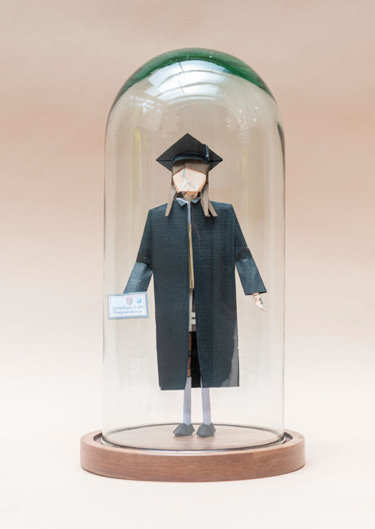 Graduada de preparatoria con toga, dentro de capelo de vidrio sobre base de madera
